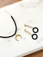 Load image into Gallery viewer, Black macrame necklace with gold crescent moon pendant and black hoop earrings on a marble surface.
