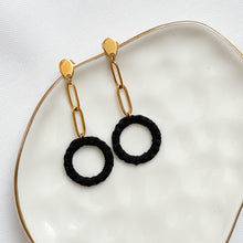 Load image into Gallery viewer, Black circular earrings with gold chains on a white plate

